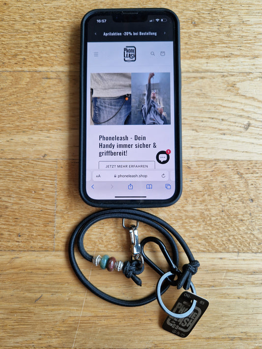 Original Phoneleash Neo Hippie mit Edelstein Indischem Achat Handykette mit elastischem Band für sicheren, stilvollen Smartphone-Gebrauch. In Deutschland gefertigt, bietet sie Langlebigkeit und ein dezentes Design mit einer Friedensbotschaft durch Peace-Zeichen und Herz auf dem Label. Darstellung mit Handy.