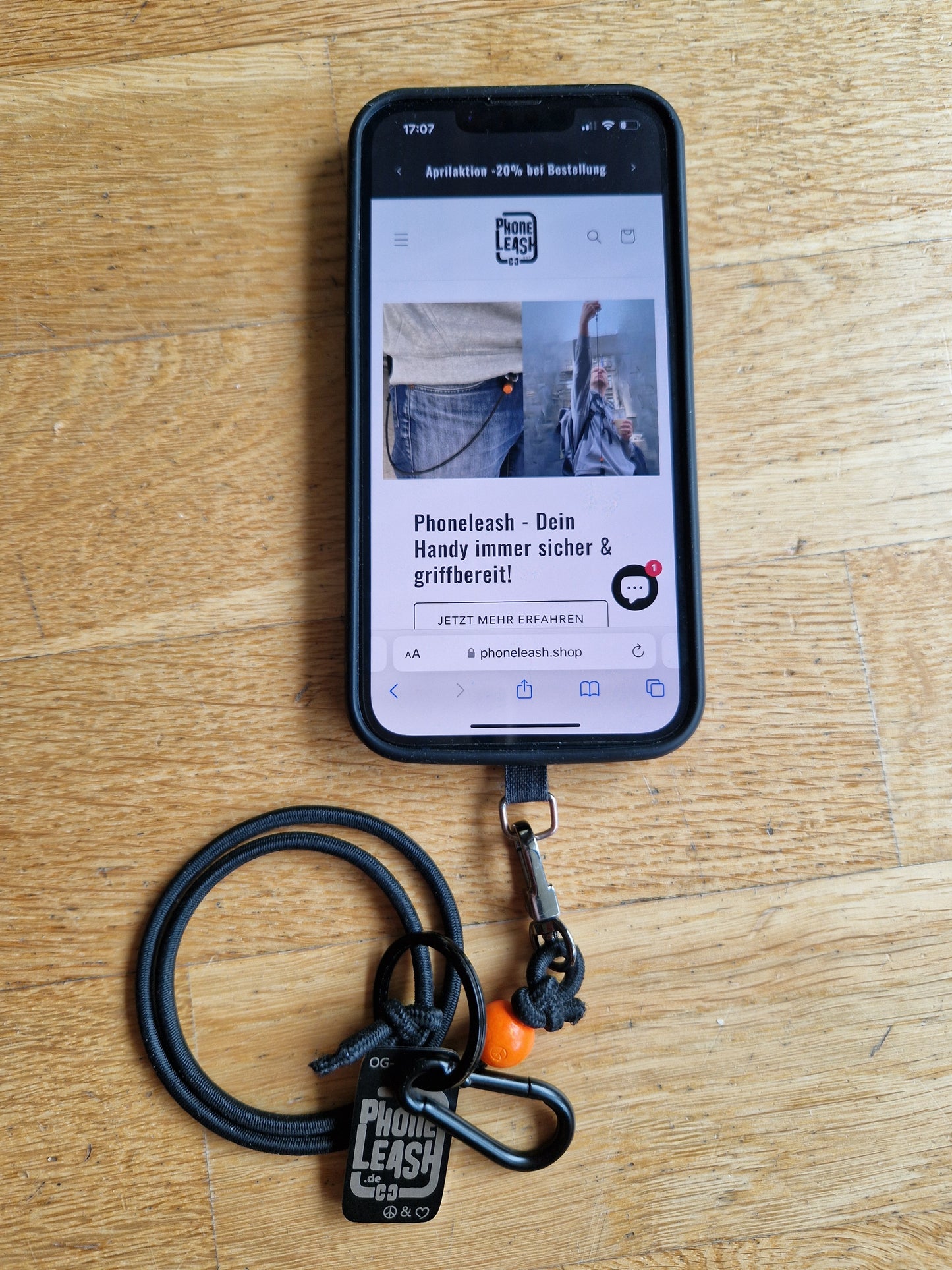 Original Phoneleash Schwarz Orange Handykette mit elastischem Band für sicheren, stilvollen Smartphone-Gebrauch. In Deutschland gefertigt, bietet sie Langlebigkeit und ein dezentes Design mit einer Friedensbotschaft durch ein handgestanztes Peace-Zeichen und Herz. Darstellung mit Handy.