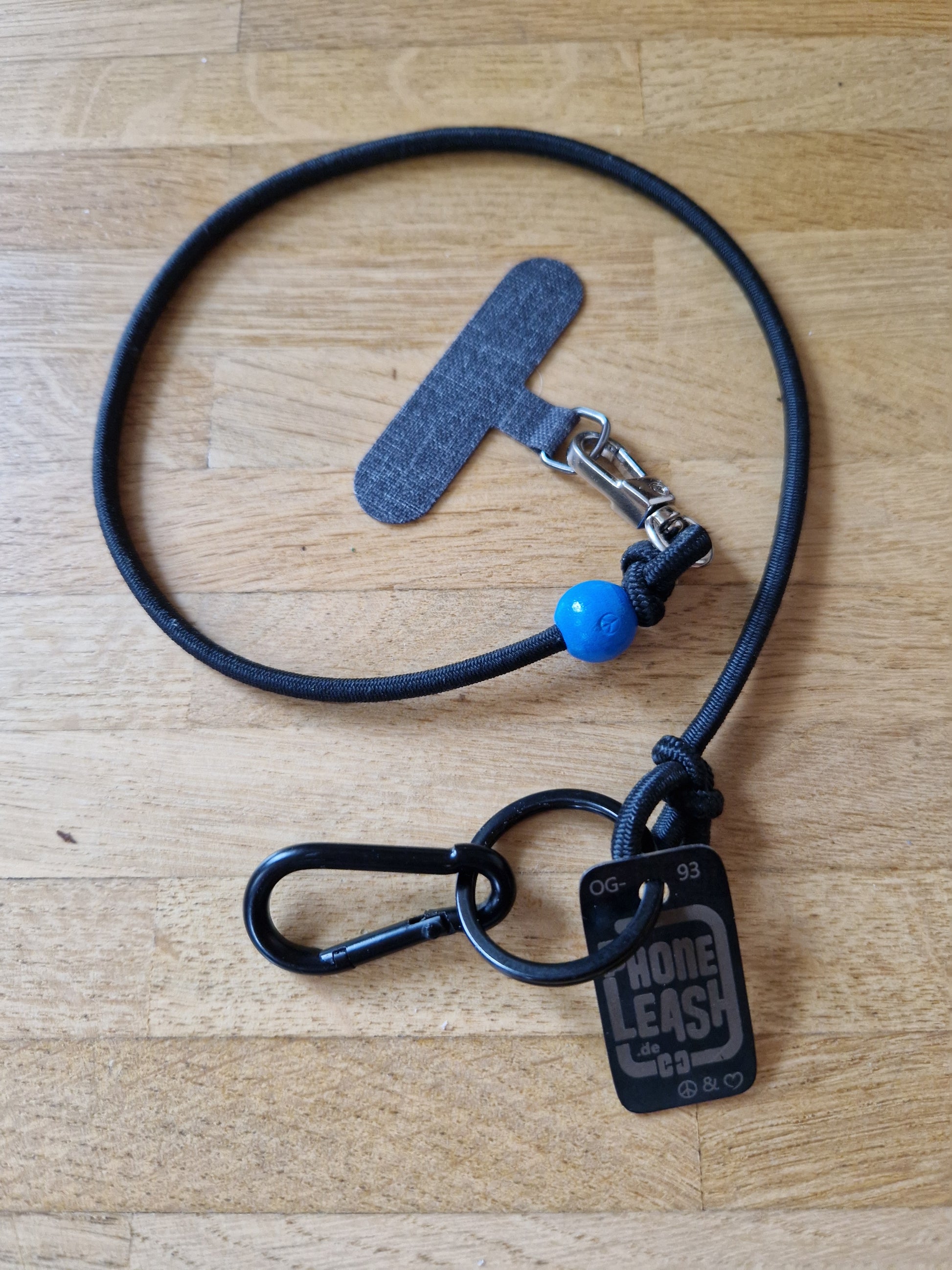 Original Phoneleash Schwarz Blau Handykette mit elastischem Band für sicheren, stilvollen Smartphone-Gebrauch. In Deutschland gefertigt, bietet sie Langlebigkeit und ein dezentes Design mit einer Friedensbotschaft durch ein handgestanztes Peace-Zeichen und Herz. Darstellung Handyband