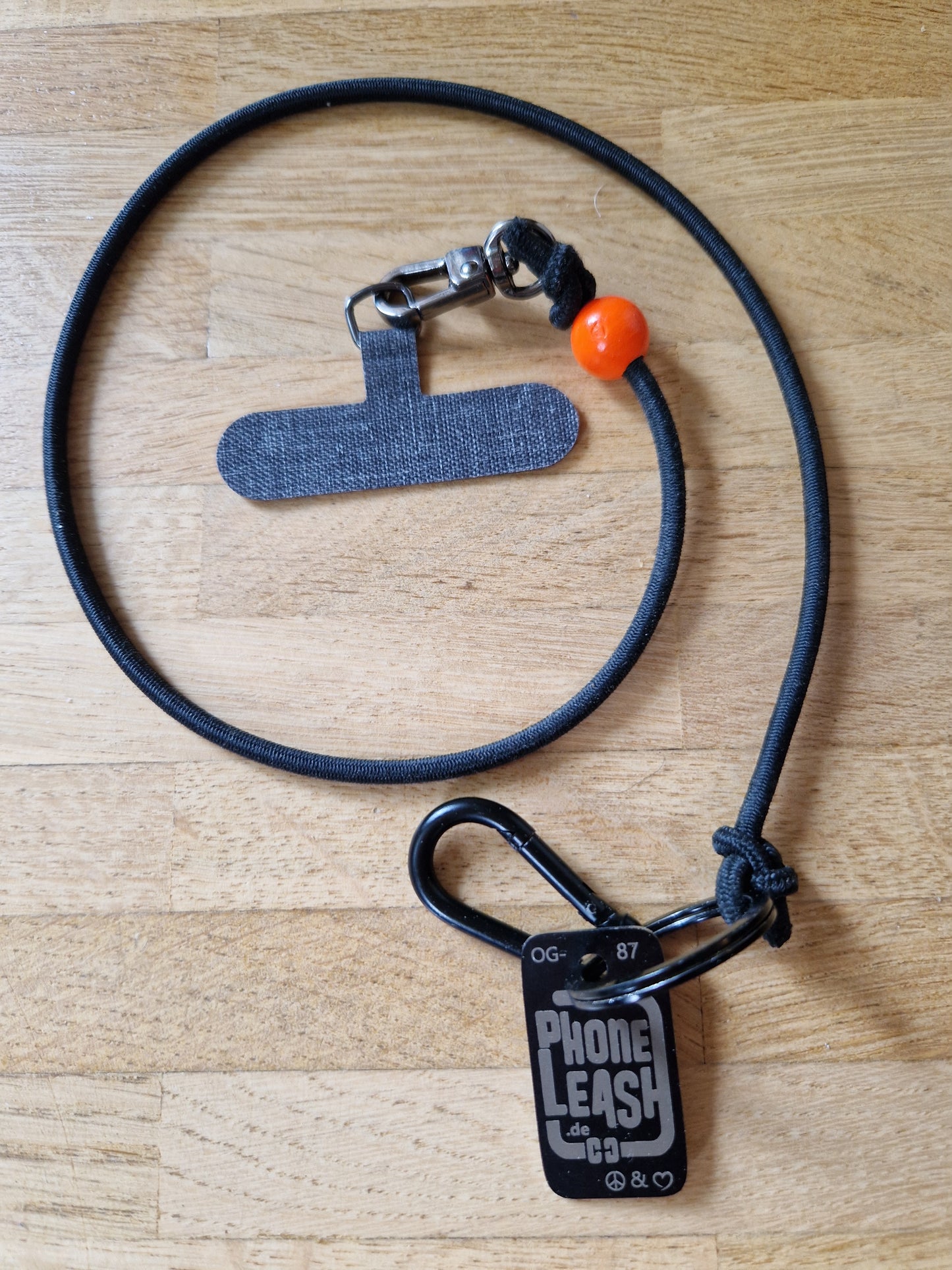 Original Phoneleash Schwarz Orange Handykette mit elastischem Band für sicheren, stilvollen Smartphone-Gebrauch. In Deutschland gefertigt, bietet sie Langlebigkeit und ein dezentes Design mit einer Friedensbotschaft durch ein handgestanztes Peace-Zeichen und Herz. Darstellung Produkt